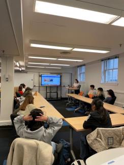 "Classroom with approximately 15 people seated around a large U-shaped table. Students are facing away from the camera looking at the instructor, who is selecting an object file to print from a website on a large screen"
