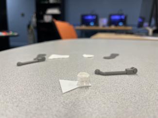 "a table on which three flat white triangular objects and three grey arm-like objects are scattered. In back of the image you can see two black square 3D printers with blue lights"